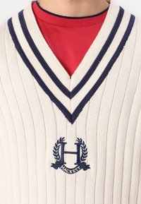 Kremfarget strikket genser med ribbestruktur, marineblå stripe ved V-halsen og brodert logo med "HACKETT" og laurbærdetaljer.