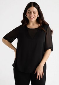 Blusa nera con maniche corte trasparenti, scollatura a V e vestibilità ampia. Il tessuto ha una texture liscia e dettagli minimi.