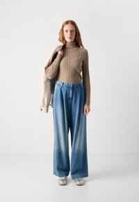 Pull en tricot beige, jeans à jambes larges bleus avec une finition délavée, et baskets argentées. Une légère veste portée sur une épaule.