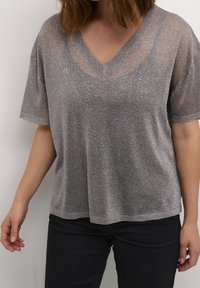 Top argento, luccicante, a maniche corte con scollatura a V e una vestibilità ampia. Tessuto morbido. Abbinato a pantaloni neri, che mostrano un look rilassato.