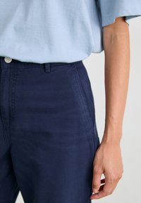 Pantalon en coton bleu marine à la texture lisse, doté d'une fermeture à bouton, de passants de ceinture et de poches latérales, associé à un haut bleu clair.