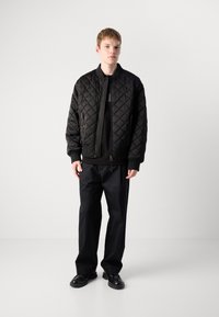 Jachetă bomber neagră, cu o quiltare, cu manșete și tiv dintr-un material ondulat, închidere cu fermoar, purtată deasupra unui pullover negru și asociată cu pantaloni închiși la culoare și încălțăminte neagră.