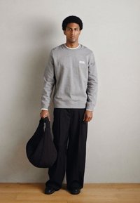 Sweatshirt cinzenta com uma pequena etiqueta branca, combinada com calças pretas largas e segurando uma bolsa preta acolchoada. Fundo simples.