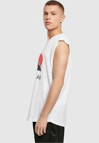 Weißes Baumwoll-Ärmelshirt ohne Ärmel mit grafischem Druck, der rote, schwarze und graue Elemente enthält, und einem lockeren Schnitt, präsentiert an einem Model.