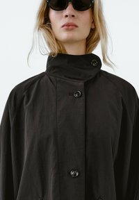 Černá knoflíková bunda s vysokým límcem, vyrobená z texturované látky a lesklých knoflíků. Model má na sobě tmavé sluneční brýle.