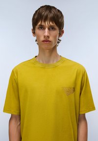 Gul bomulds T-shirt med rund hals og korte ærmer. Har et lille brunt grafisk logo på venstre brystområde. Glat tekstur.