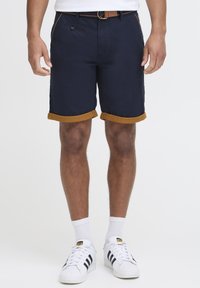 Mørkeblå shorts med rullede beige opplegg, sidelommer og frontknappelukking. Kombinert med hvite sneakers og hvite sokker.