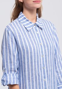 Blue and white striped linen shirt with a button-down collar, roll-up sleeves, and pearlescent buttons. Light texture with casual design.