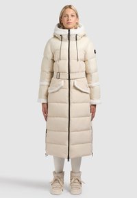 Lange donsjas in crème met een capuchon gevoerd met namaakbont, een ceintuur en afritsbare zijzakken. Gladde, doorgestikte textuur met contrasterende accenten.