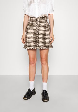 Vrouw die een rok met bruin-wit hondenbotmotief draagt, een witte blouse met pofmouwen, witte sokken en zwarte loafers met kwastjes, staand tegen een effen achtergrond.