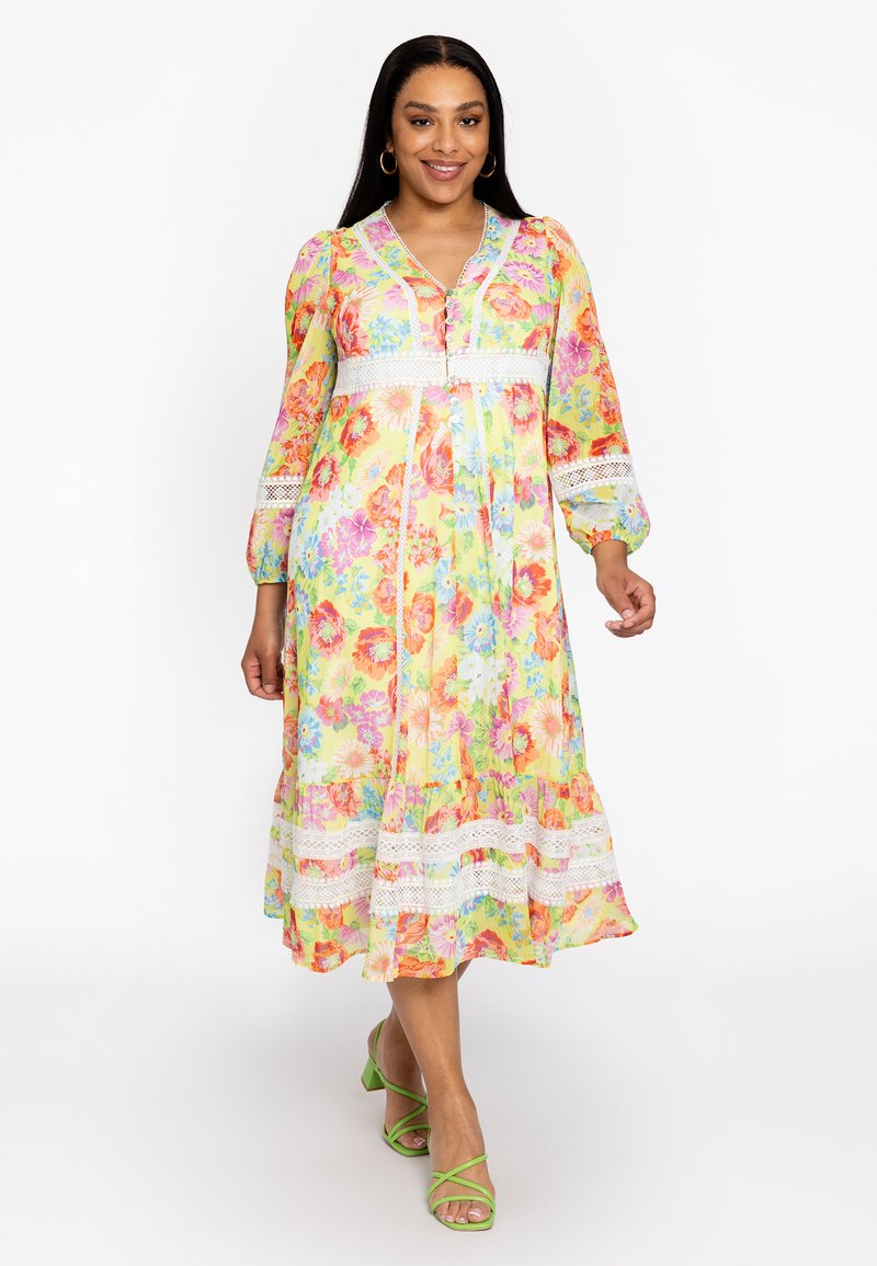 Robe maxi florale jaune, ornée de motifs colorés, de manches bouffantes, de dentelle et de détails de boutons. Associée à des sandales à lanières vertes.