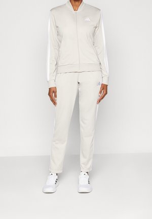 Person trägt eine hellbeige Adidas-Trainingsanzug mit weißen Streifen an Ärmeln und Hosen, kombiniert mit weißen Adidas-Sneakern.