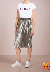 Camiseta blanca con "HUGO" en burdeos, combinada con una falda midi de metalizada plata con pliegues y zapatillas blancas. La falda cuenta con una cinturilla elástica gris.