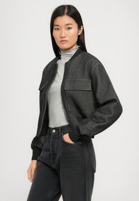 Veste bomber noire courte avec deux poches avant, poignets côtelés et fermeture éclair, portée sur un col roulé gris et un jean noir taille haute.