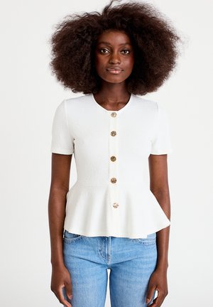 Donna con capelli ricci e voluminosi che indossa un top bianco a maniche corte con baschina e bottoni effetto tartaruga, e jeans blu, in piedi davanti a uno sfondo semplice.