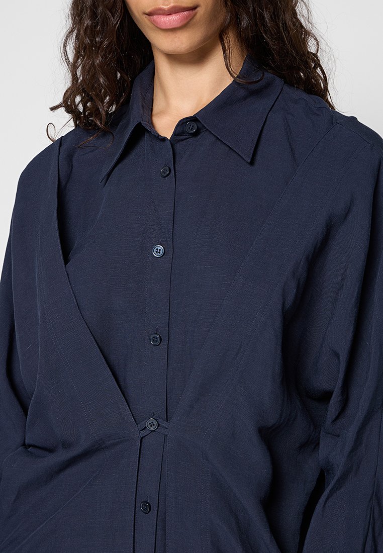 Camicia blu navy con bottoni, dotata di un colletto strutturato, realizzata in un tessuto morbido, con una vestibilità rilassata e un particolare dettaglio annodato in vita.