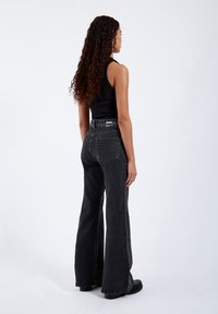 Vrouw met lang krullend haar, die een zwart mouwloos topje en zwarte high-waisted wijde jeans draagt, staand zijwaarts tegen een witte achtergrond.