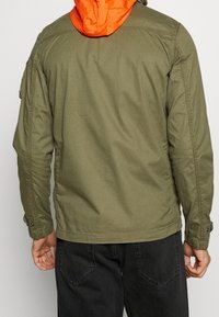 Olivgrön jacka med orange huva. Har en avslappnad passform, sidfickor och ärmuppslag med knappar. Tillverkad av lätt tyg.