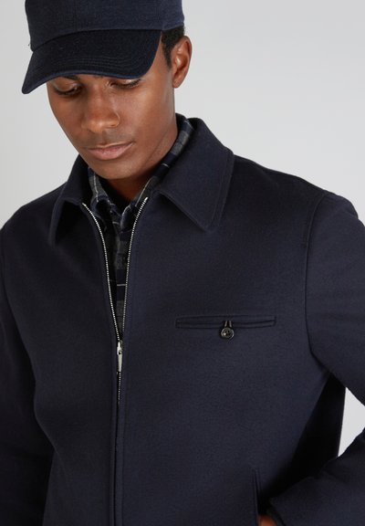 Marineblauer Wollmischungsjacke mit Reißverschluss, spitzem Kragen und Brusttasche. Tragen über einem karierten Hemd. Abgerundet mit einer marineblauen Mütze.