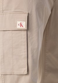 Beige stof cargo broeken met een zijzak. Heeft een opvallend CK-logo label in het rood op een witte achtergrond. Gladde textuur en regular fit.