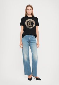 Schwarzes T-Shirt mit einem goldenen runden Grafiklogo, kombiniert mit hellblauen weit geschnittenen Jeans und schwarzen High Heels.
