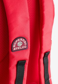 Röd ryggsäck med en texturerad yta, utrustad med vadderade remmar. Svarta detaljer och en cirkulär logotyp med texten "SUPERDRY ATHL.CO."