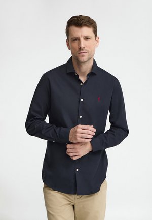 Ein Mann trägt ein marineblaues Hemd mit Knöpfen und einem kleinen roten Logo auf der Brust sowie beige Hosen und richtet seinen linken Ärmel.