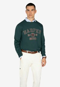 Felpa verde scuro con grafica "HARPER 1985", indossata sopra una camicia a colletto azzurra. Abbinata a pantaloni cargo beige e cintura marrone.
