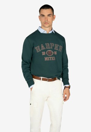 Sudadera verde oscura con el gráfico "HARPER 1985", usada sobre una camisa de color azul claro con cuello. Combinada con pantalones cargo color crema y un cinturón marrón.