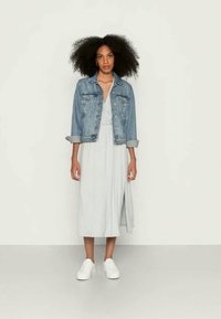 Vestido midi cinza claro com um design envolvente, combinado com uma jaqueta de denim azul, com dois bolsos frontais e mangas enroladas. Ténis brancos completam o visual.