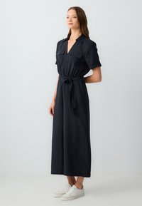 Robe bleu marine à manches courtes avec un col, fermeture éclair, deux poches poitrine et une ceinture à la taille ; associée à des baskets blanches.