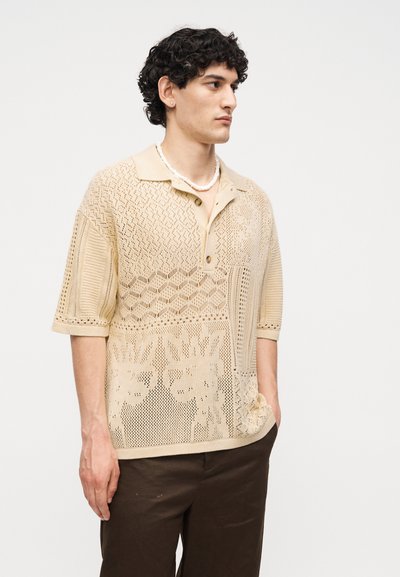 Άνδρας με κατσαρά μαλλιά που φοράει μπεζ πλεκτό polo με κοντά μανίκια και σχέδια και σκούρο καφέ παντελόνι, κοιτάζει αριστερά με το ένα χέρι στην τσέπη.