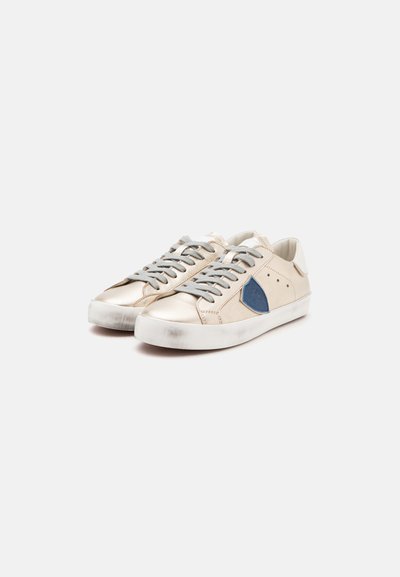 Ζευγάρι χαμηλά sneakers σε εκρού χρώμα με γκρι κορδόνια, λευκές σόλες και μπλε ετικέτα σε σχήμα ασπίδας στην εξωτερική πλευρά κάθε παπουτσιού.