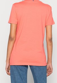 Osoba ubrana w koralową koszulę z krótkim rękawem i niebieskie dżinsy, pokazana od tyłu na tle jasnego, jednolitego tła.