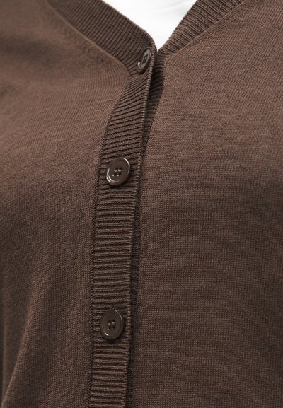 Κοντινή λήψη καφέ πλεκτής ζακέτας με ριμπ λεπτομέρεια και τρία σκούρα κουμπιά κλειδωμένα πάνω από λευκό εσωτερικό ένδυμα.