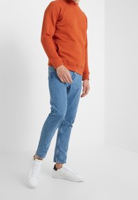 Pomarańczowa bluza z raglanowymi rękawami, zestawiona z jasnoniebieskimi jeansami i białymi tenisówkami z czarnymi akcentami, na białym tle.