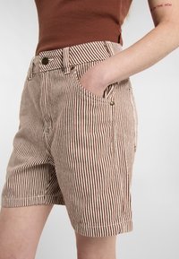 Pantalones cortos a rayas en marrón y crema con bolsillos laterales, cierre frontal de botón y una cinturilla ajustada. Hechos de un tejido texturizado.