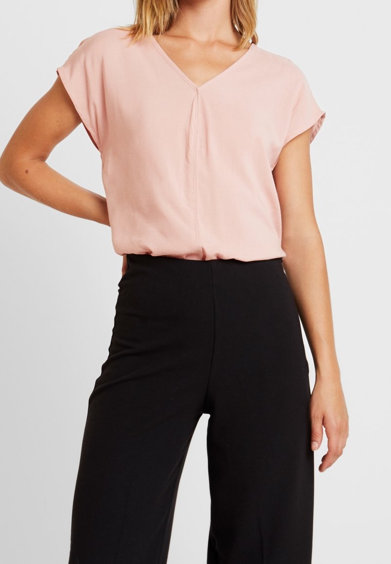 Femme portant un chemisier rose clair à manches courtes et col en V, rentré dans un pantalon noir taille haute à jambes larges, une main sur la hanche.