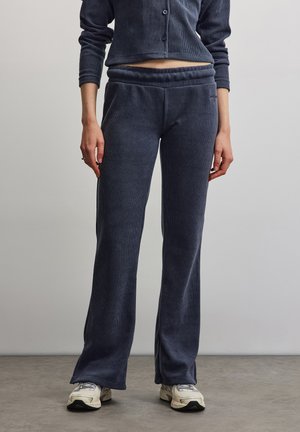 Persoon die donkergrijze geribde wijde broek en crèmekleurige sneakers draagt, staand op een grijze vloer tegen een effen lichte achtergrond.