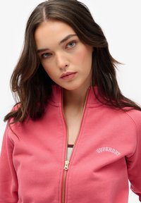 Sudadera con cremallera rosa hecha de forro polar suave, con cremallera dorada y logotipo blanco de "SUPERDRY" en el pecho izquierdo. Textura suave, ajuste casual.