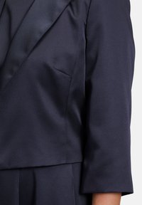 Granatowa marynarka z satynowymi klapami, usztywniony fason, rękawy do łokcia oraz tekstura z ukośnymi żebrami. Detal kieszeni z przodu.