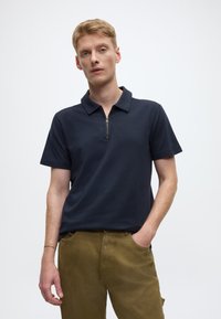 Granatowa koszulka polo z zamkiem błyskawicznym, teksturowana tkanina, krótkie rękawy, noszona z beżowymi spodniami khaki z bocznymi kieszeniami o luźnym kroju.