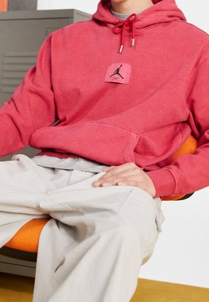 Osoba w czerwonej bluzie z kapturem z czarnym logo Jumpman i beżowych spodniach siedzi na pomarańczowym krześle, a w tle znajduje się szara szafka.