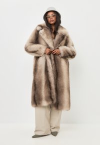 Kožich z umělé kožešiny v odstínech světlé hnědé s tmavšími pruhy, s velkým límcem a širokými rukávy, doplněný béžovými kalhotami a špičatými zelenými botami.