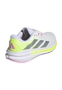 Zapatilla deportiva blanca con parte superior de malla texturizada, acentos en neón amarillo, rayas grises y una suela blanca con un acento rosa.