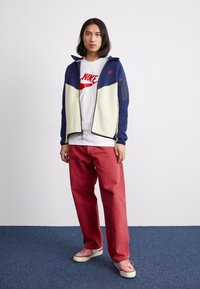 Herre zip-jakke med marineblå og cremefarvede farveblokke, parret med røde bukser og pink sneakers. Har et logo på skjorten og en lynlåslomme foran.