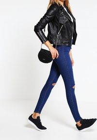 Giacca da motociclista in pelle nera, jeans skinny strappati blu, scarpe slip-on nere e piccola borsa a tracolla nera. Sfondo neutro.