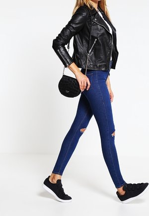 Czarna skórzana kurtka motocyklowa, niebieskie przetarte skinny jeansy, czarne buty slip-on i mała czarna torba na ramię. Neutralne tło.