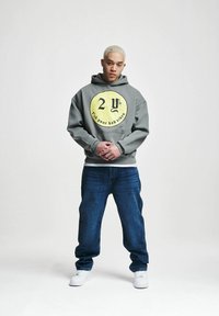 Grå hettegenser med gul sirkulær grafikk som inneholder tekst. Bært med blå jeans og hvite joggesko. Modellen står med hendene sammenfoldet.