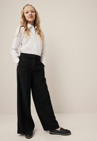 Schwarze weit geschnittene Hose mit seitlichen Taschen, kombiniert mit einem weißen Hemd mit Knöpfen. Das Model trägt schwarze Slipper mit strukturierter Oberfläche.